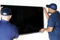 Dois profissionais posicionam uma TV grande na parede durante instalação de suporte de TV.