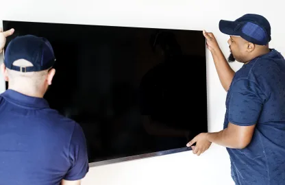 Dois profissionais posicionam uma TV grande na parede durante instalação de suporte de TV.