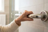Mão de pessoa idosa segurando barra de apoio em inox fixada na parede de azulejos do banheiro após instalação de barra de apoio para idosos.